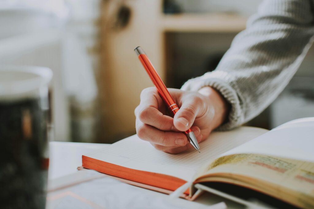 A person writing in a notebook with a red ballpoint pen, showcasing detail and focus.